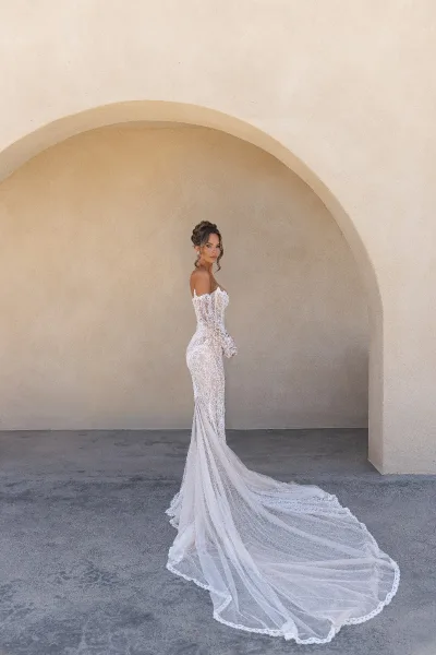 Bridal portrait of a bride in lace gown beneath a stucco arch, highlighting off-the-shoulder sleeves.