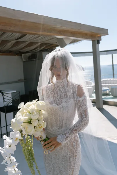 Bridal portrait of a bride with veil on a rooftop terrace overlooking the ocean, framed by a glass railing and modern lounge seating.