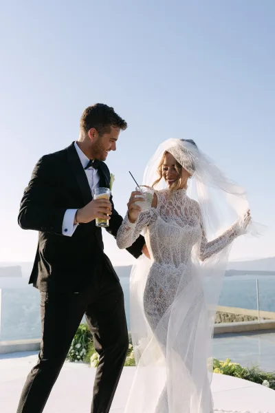 Couple portrait of a bride and groom celebrating, her bridal veil trailing, on a terrace above the ocean under a clear blue sky.