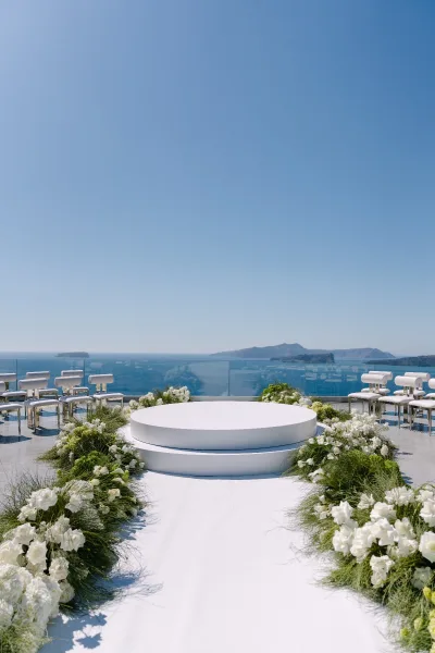 Ceremony setup featuring refined outdoor ceremony decor on a terrace, with a round platform and ocean view framed by a glass railing.