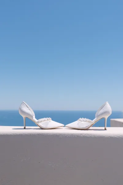 Bridal shoes, pearl wedding heels with a pointed toe, set against a white wall with blue sky and an ocean horizon softly blurred behind.