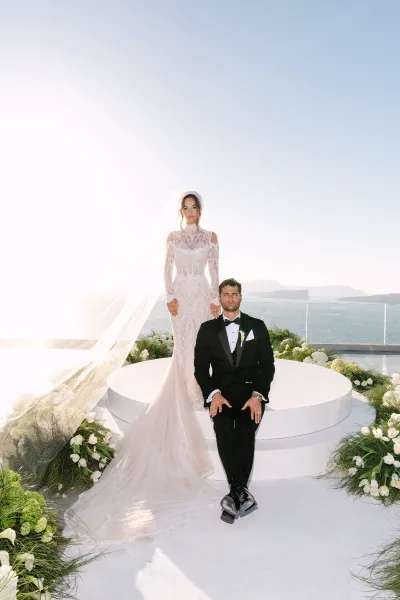 Couple portrait of a bride and groom in a formal pose on a round platform, framed by ocean views and coastline cliffs behind a glass railing.