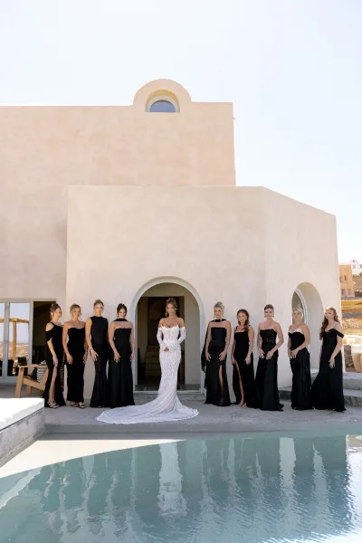 Bride and bridesmaids in black bridesmaid dresses pose by a stucco arched doorway beside a swimming pool beneath a clear sky.