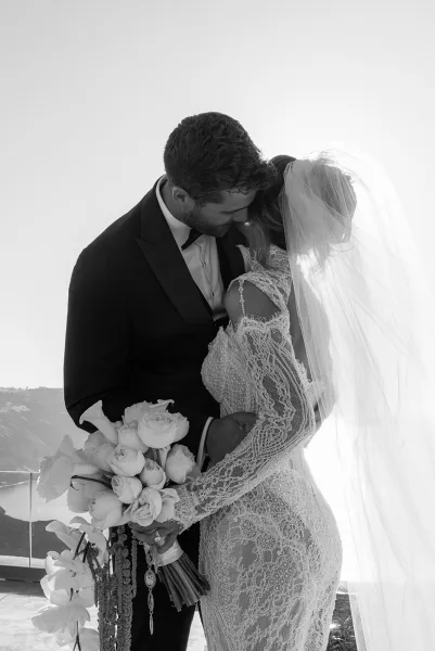 Couple portrait of a bride and groom embrace, holding a bridal bouquet, by ocean cliffs behind a glass railing under the sky.
