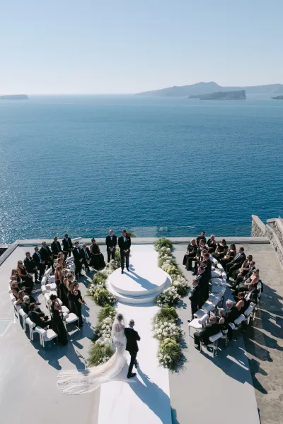 Wedding processional with a bride walking down the aisle on a white aisle runner, set on an oceanfront terrace with blue sky and distant islands.