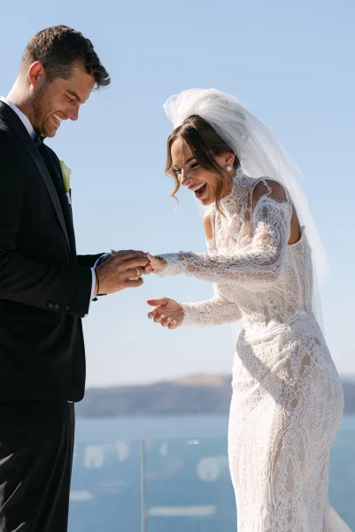 Wedding ring exchange at a seaside ceremony moment as the groom slides the band onto her finger, bride in a tulle veil, ocean and cliffs beyond.