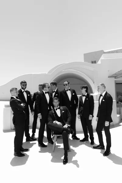 Groomsmen portrait in black tie groomsmen style, lined up by white stucco walls and an arched doorway, one wearing sunglasses.