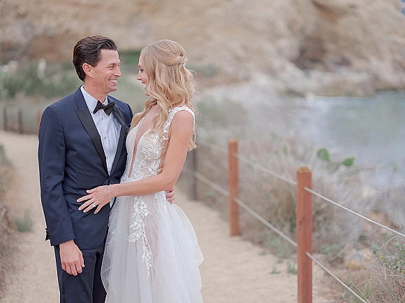 Couple portrait of bride and groom portrait hugging on a sandy coastal path by ocean cliffs, lace wedding dress and tuxedo visible