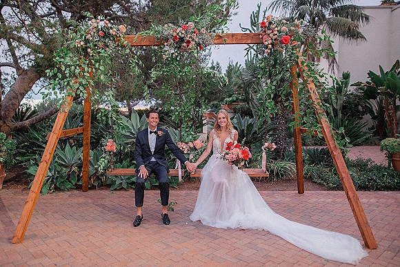 Couple portrait of bride and groom holding hands on a wooden swing with floral garland in a garden patio with palm trees and succulents