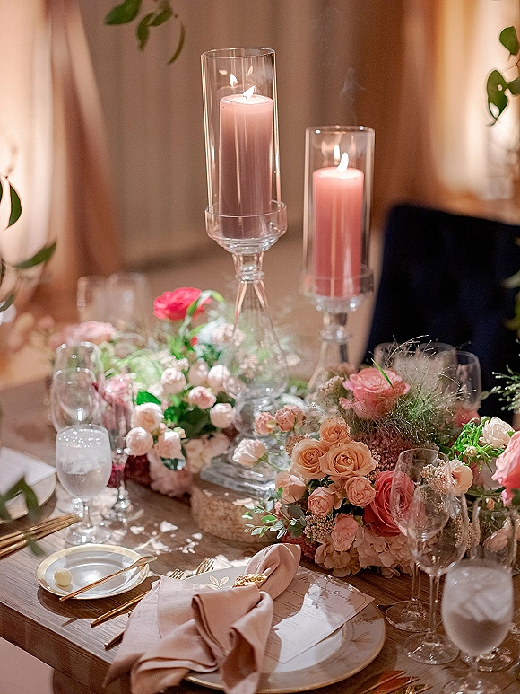 Reception tablescape with a wedding table centerpiece of roses and greenery, pink taper candles in glass hurricanes, and gold flatware before draped fabric backdrop