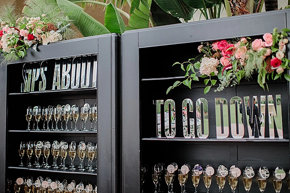 Wedding champagne wall with champagne flutes and pre-poured champagne, mirrored letter sign, and floral greenery accents on a lawn with tropical leaves