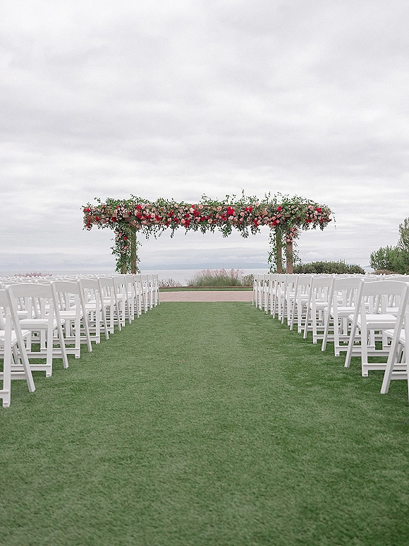 Ceremony setup with a floral arch and greenery garland, white folding chairs lining an aisle on a grass lawn with ocean view under cloudy sky