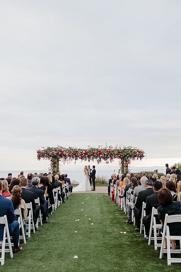 Outdoor wedding ceremony with a floral arch as bride and groom stand at the altar, ocean coastline backdrop and white aisle chairs