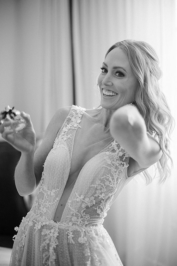 Bridal portrait of a smiling bride in a lace wedding dress with deep V neckline, wavy half-up hair and earrings by window light indoors