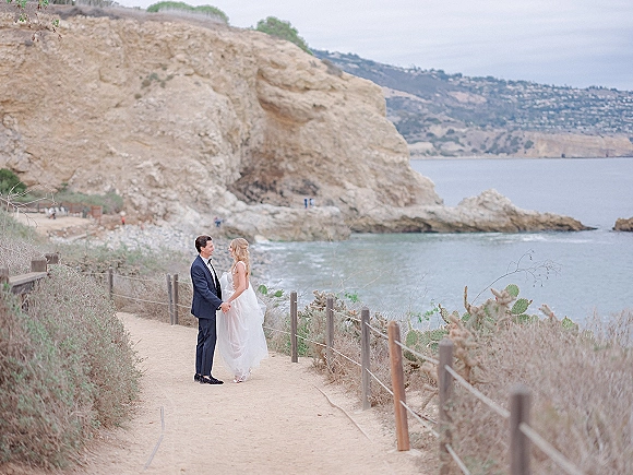 Couple portrait of bride and groom holding hands on coastal cliffs, her veil blowing as ocean waves roll under an overcast sky