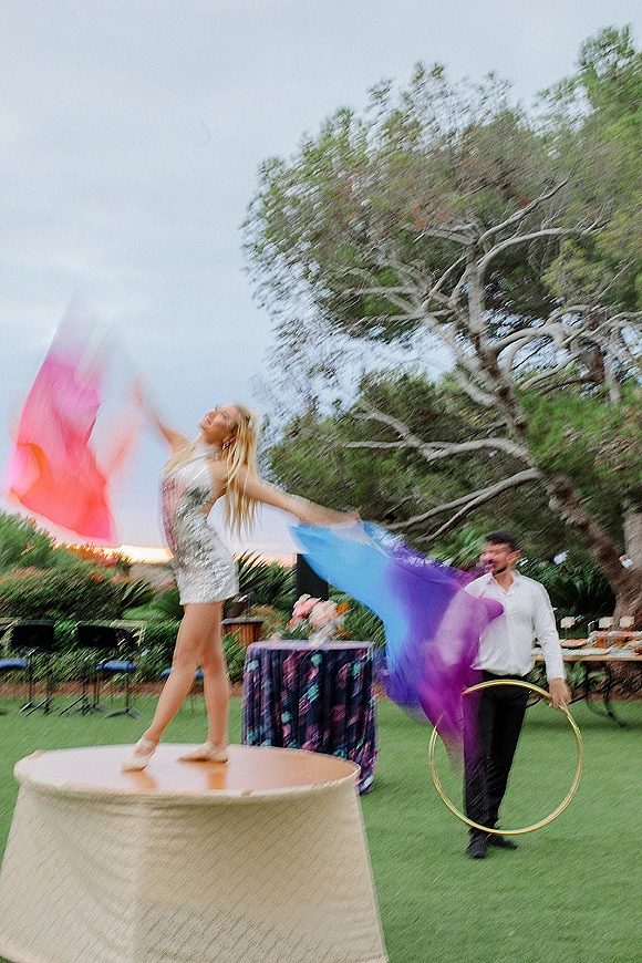 Wedding entertainment with a hula hoop performer twirling colorful fabric flags on an outdoor lawn, with cocktail tables and trees behind