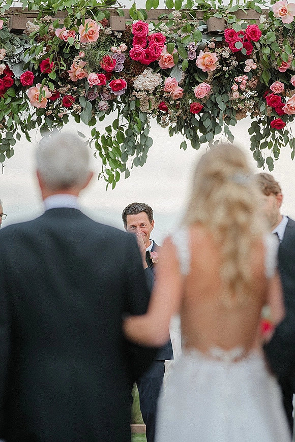 Wedding processional as bride walking down aisle with long veil toward groom under wooden pergola floral arch, sky and landscape behind