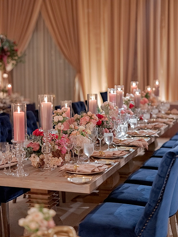 Reception tablescape with long head table decor featuring blush roses, greenery, tall cylinder candles, gold flatware and navy velvet chairs under draped uplighting