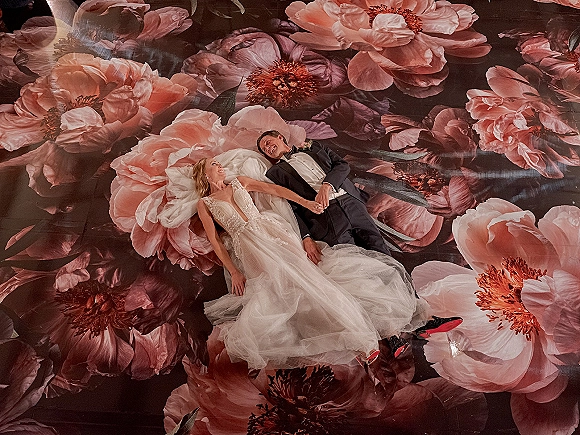 Couple portrait in an overhead wedding photo, bride and groom lying on a floral patterned floor, veil spread, tux and red-sole heels visible