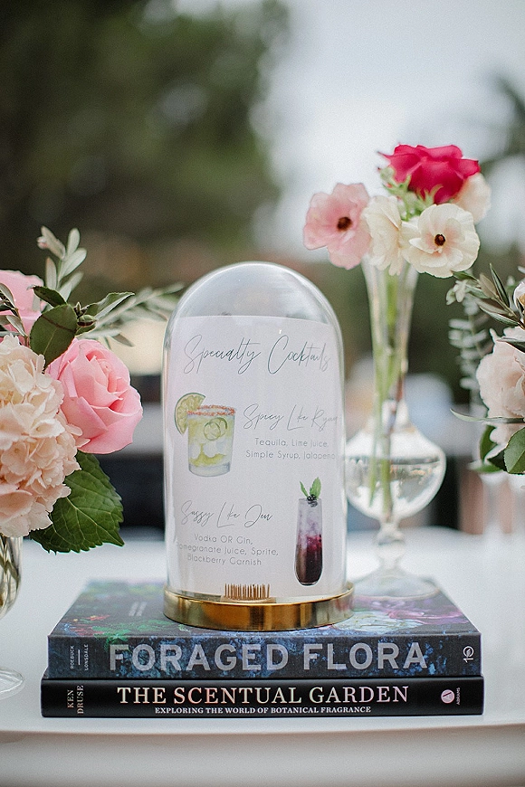 Wedding cocktail menu acrylic sign on a brass stand with pink roses and hydrangea on stacked books, greenery and trees blurred behind