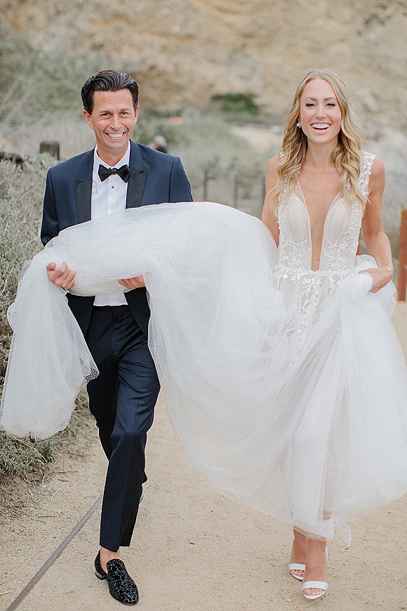Couple portrait of bride and groom walking as he lifts her dress train, smiling on an outdoor hillside path with shrubs, her tulle skirt flowing