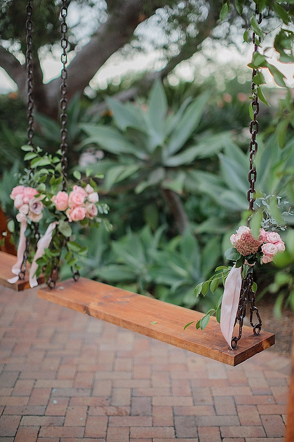 Wedding swing decor with pink roses and greenery on metal chains, ribbon streamers on a wood seat, set amid garden foliage over brick patio
