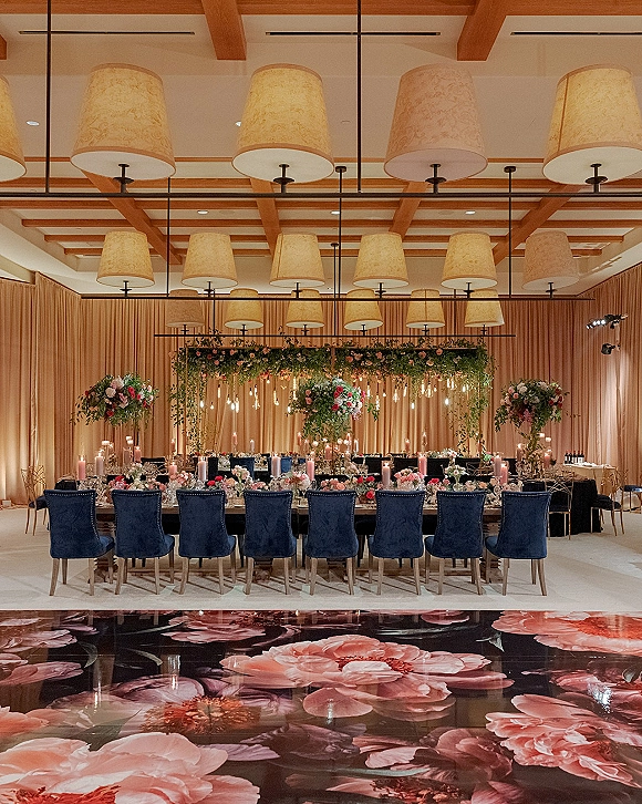 Reception tablescape with head table decor on a long banquet table, blue velvet chairs, candlelit florals, and hanging greenery in a wood-beam ballroom