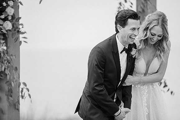 Wedding couple portrait in black and white, bride and groom laughing hand in hand beside a white wall with foliage shadows