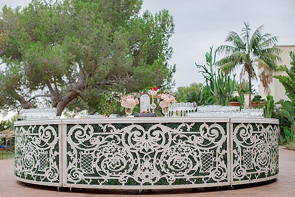 Wedding bar setup with ornate white carved bar front, champagne flutes and blush florals, set on an outdoor patio with palms and cactus