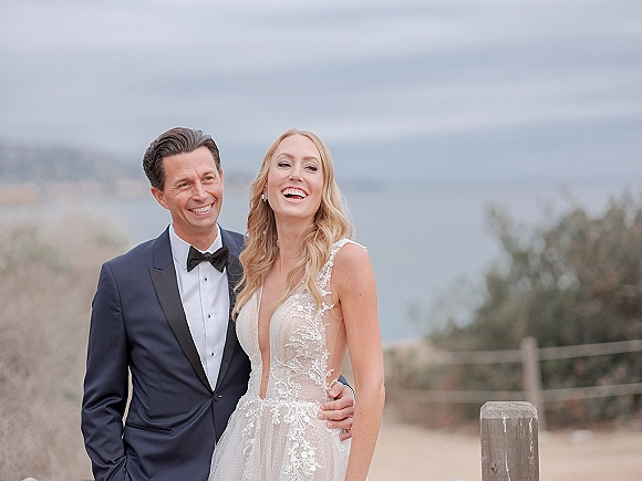 Couple portrait of bride and groom laughing on a beach, her lace wedding dress flowing as he holds her waist by sand dunes and fence