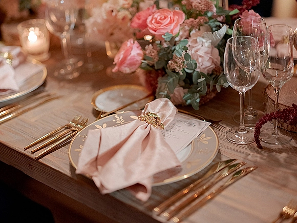 Reception tablescape with pink floral centerpiece, blush napkins, gold flatware, patterned plates, menu card, and votive candles on a wood table