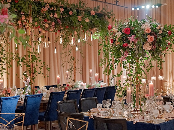 Reception tablescape with wedding head table decor, overhead floral installation and taper candles beneath string lights and a draped wall backdrop