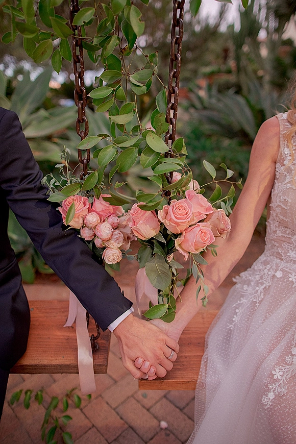 Wedding bouquet of pink roses and greenery with long ribbon held by bride in lace dress beside groom on a wooden garden swing over brick path
