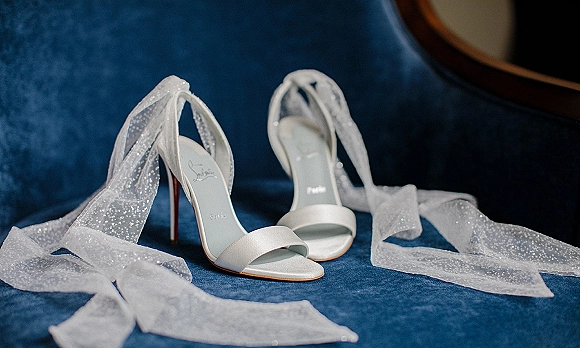 Bridal shoes, white wedding heels with glitter tulle ribbon ankle ties and stiletto heels displayed on a blue velvet sofa by a round mirror