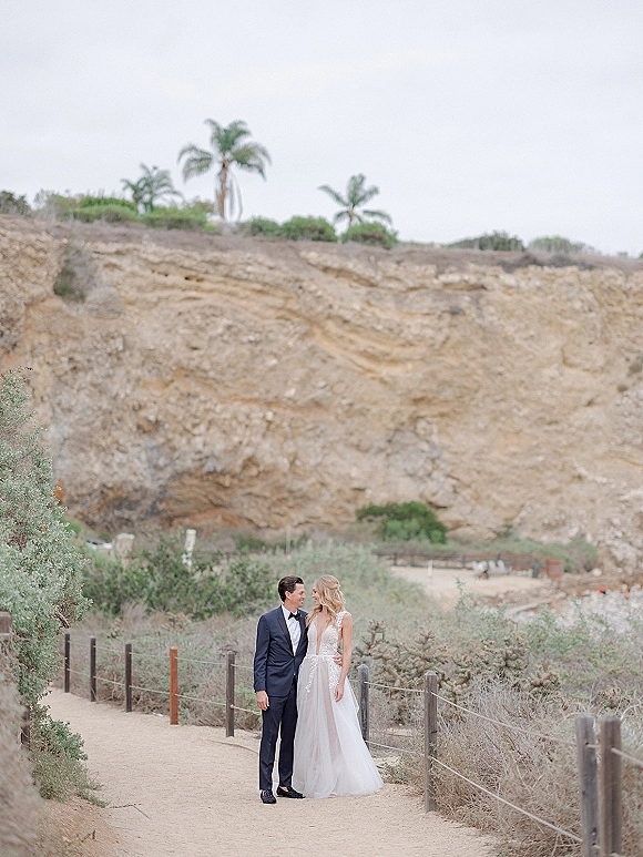 Couple portrait of bride and groom outdoors on a sandy path by a wooden fence, bride in a lace wedding dress, under overcast sky