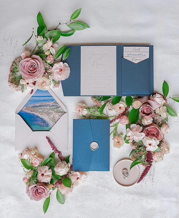 Invitation suite with blue wedding invitations in a folio, wax seal, RSVP and accommodation cards, styled with pink roses on white texture