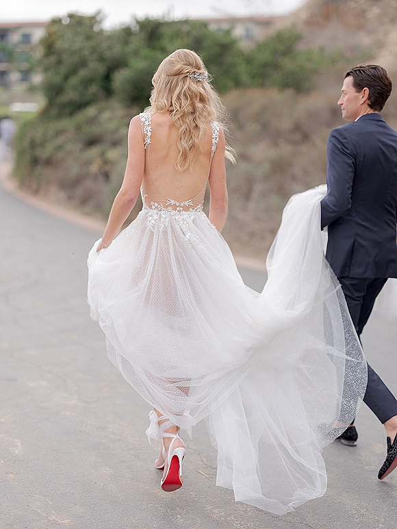 Wedding couple portrait of bride and groom walking away holding hands on a paved hillside road, her backless gown train flowing behind