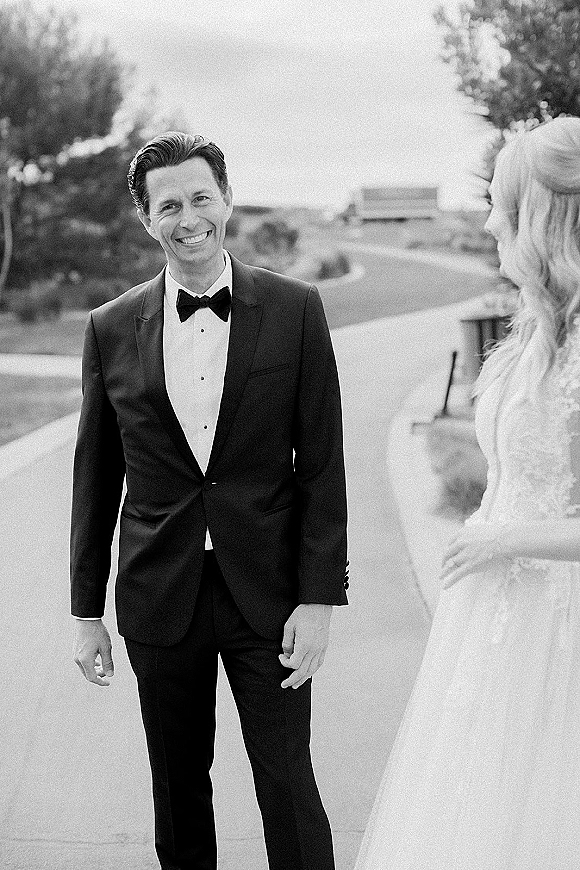 Groom portrait in a black tuxedo, bow tie and dress shirt, smiling beside the bride’s lace sleeves on an outdoor terrace with stone wall and hills