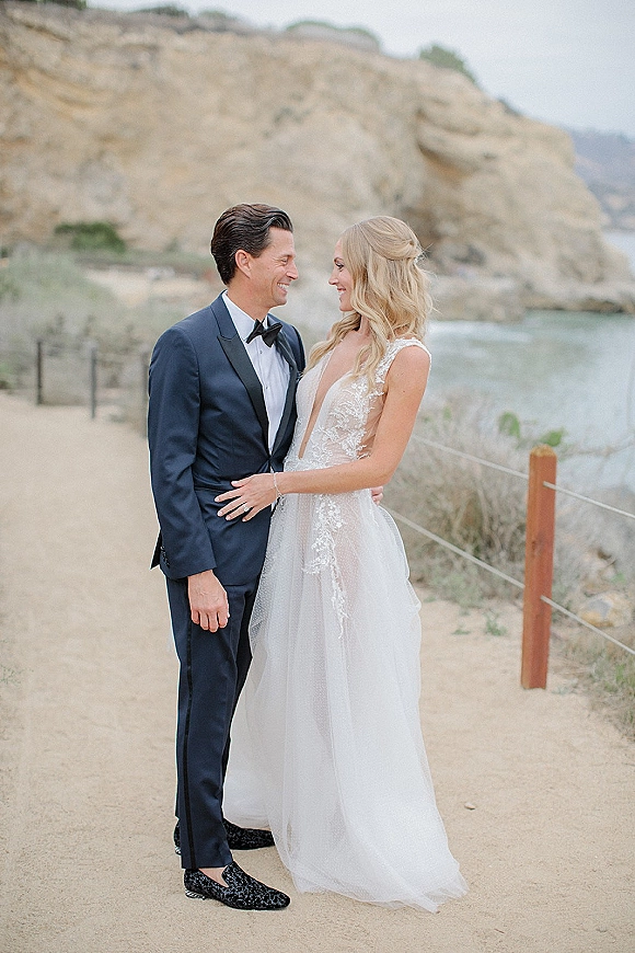 Couple portrait of bride and groom embrace, bride in lace deep V gown and groom in tuxedo on coastal cliffs by the ocean