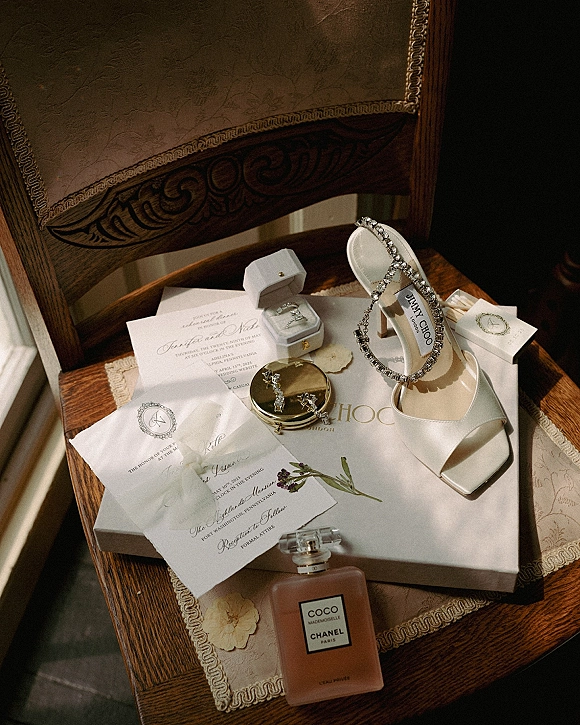 Wedding details flatlay with wedding invitation suite, rings in boxes, earrings, heels, perfume, ribbon and wax seal on a lace runner in window light