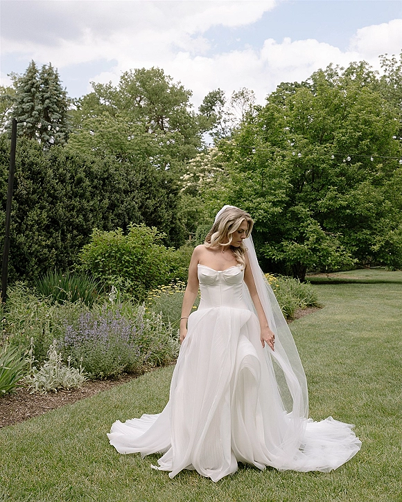 Bridal portrait of a bride in a strapless wedding dress with a long veil and train, looking down on a garden lawn under string lights