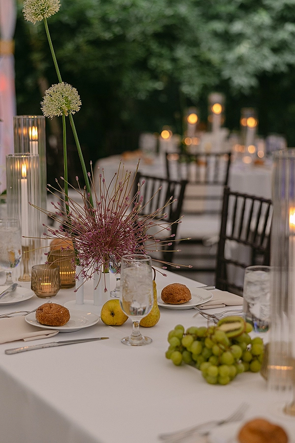 Reception tablescape with outdoor reception table styling, tall allium centerpiece in white vases with candles, fruit, and black chairs under bistro lights