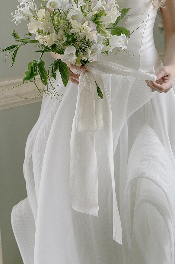 Bridal bouquet of white flowers and greenery with a long ribbon, held against a pleated bridal gown with engagement ring visible