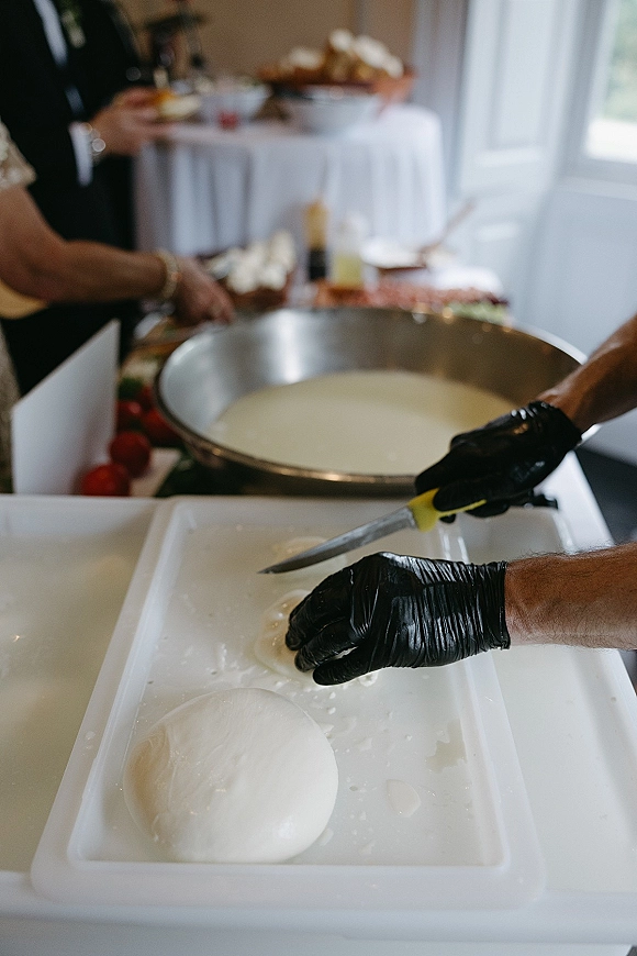 Wedding food station with chef in black gloves slicing fresh mozzarella beside tomatoes and bowls on a buffet table by windows