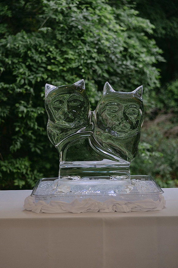 Ice sculpture wedding ice sculpture of two cats on a serving tray with ice chips on a white tablecloth, set against green foliage