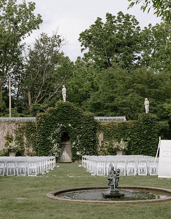 Ceremony setup with outdoor wedding ceremony seating of white folding chairs facing a fountain, with florals and ivy-covered wall backdrop