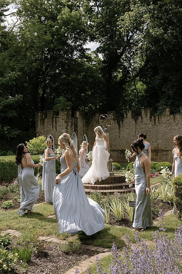 Bridesmaid group with bride with bridesmaids walking together holding white and green bouquets in light blue dresses by a stone wall garden sundial
