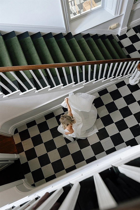 Bridal portrait of a bride twirling her strapless pleated wedding dress on a staircase with green runner and checkered floor in natural light