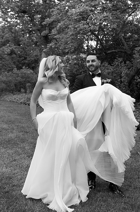 Wedding couple portrait in black and white, bride in strapless dress and veil walking with groom in tuxedo under garden string lights