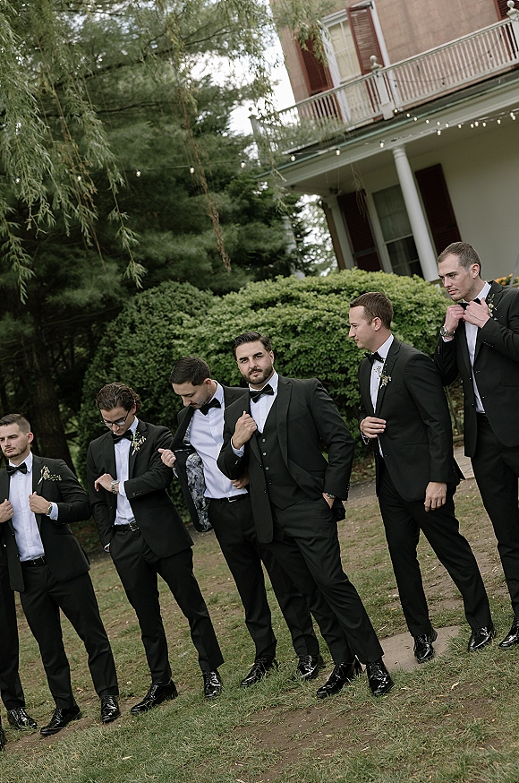 Groomsmen portrait of men in black tuxedos with bow ties and boutonnieres, standing on a lawn by shrubs and string-lit porch